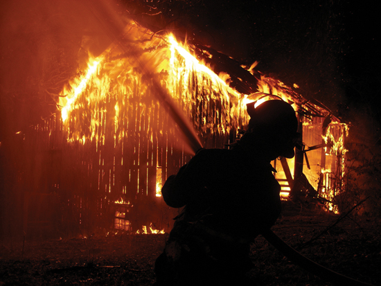 Firefighter spraying water from hose on fully envolved barn fire. Night time shot.